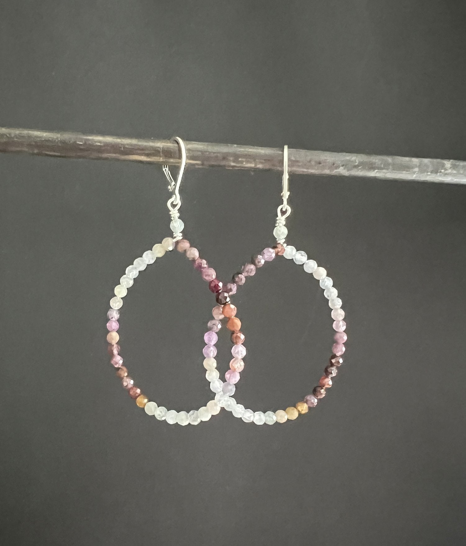 The ANN LIGHTFOOT - VARIED SPINEL HOOP EARRINGS showcase small, multicolored beads in hues of pink, purple, and orange. They hang gracefully from sterling silver earwires and are beautifully set against a dark gray background.