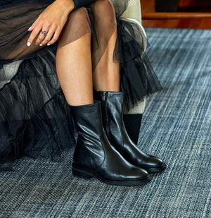 A side view of the CORDANI Blaize Leather Bootie with Side Zip, showcasing its sleek, black leather above-the-ankle design with a smooth finish. The boot boasts a mid-calf height, rounded toe, and low, chunky stacked heel. It features a convenient pull tab at the back for easy wearing. Made in Italy by CORDANI, set against a plain white background to emphasize its elegant design.