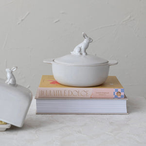 The STONEWARE BAKER WITH RABBIT FINIAL by CREATIVE COOP is a cream-colored stoneware casserole dish with side handles and a lid topped with a sitting rabbit figure as the handle.