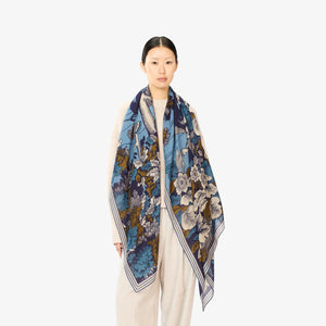 A person is wearing a beige sweater and the INOUI EDITIONS - PANTHERE SCARF, which features a blue, brown, and white floral pattern. The scarf made of washable wool is wrapped around their neck. The plain white background emphasizes the scarf and sweater, with the individual's face partially visible.