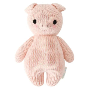 A pink knitted baby piglet, crafted from 100% cotton yarn, is shown from the back. It has small round ears, arms, a tail, and a tag attached to its side. The piglet boasts a simple, cuddly design and radiates the charm of hand-knit baby animals while supporting fair trade income. This delightful creation is known as the CUDDLE + KIND - HANDMADE BABY PIGLET by CUDDLE & KIND.