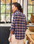 A person with wavy hair, dressed in a relaxed fit blue and red plaid Eileen shirt from Frank & Eileen, stands in a kitchen and glances over their shoulder. The kitchen features large windows, dark cabinets, and a wooden countertop with a bowl of fruit in the background.