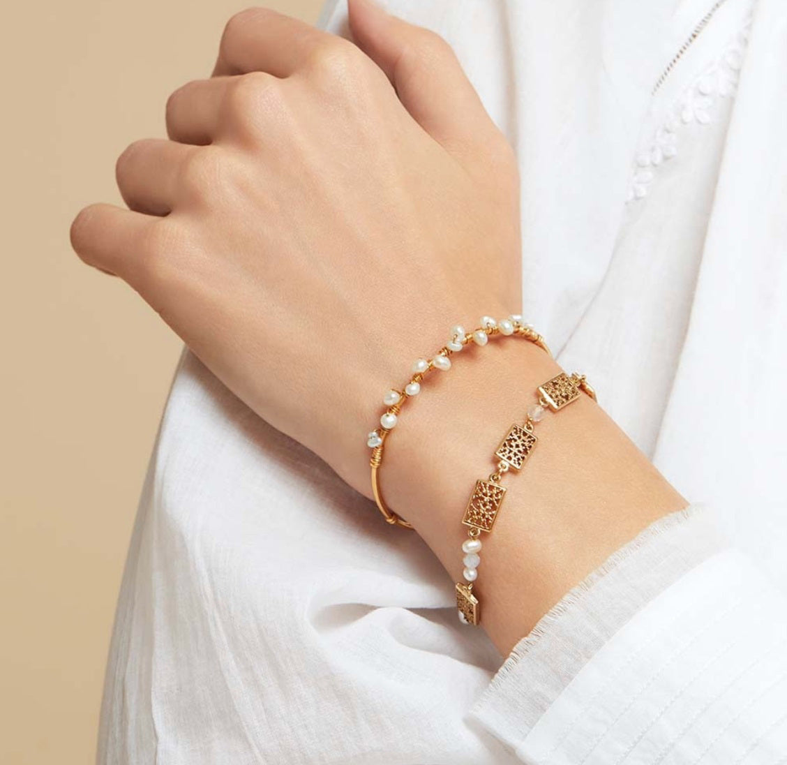 A hand with a white sleeve is adorned with two bracelets. One bracelet features small pearls, while the other is the exquisite GAS BIJOUX - CALLIOPE BRACELET, a stunning gold-plated bangle showcasing intricate geometric designs. Both pieces exude elegance and sophistication against a neutral beige background.