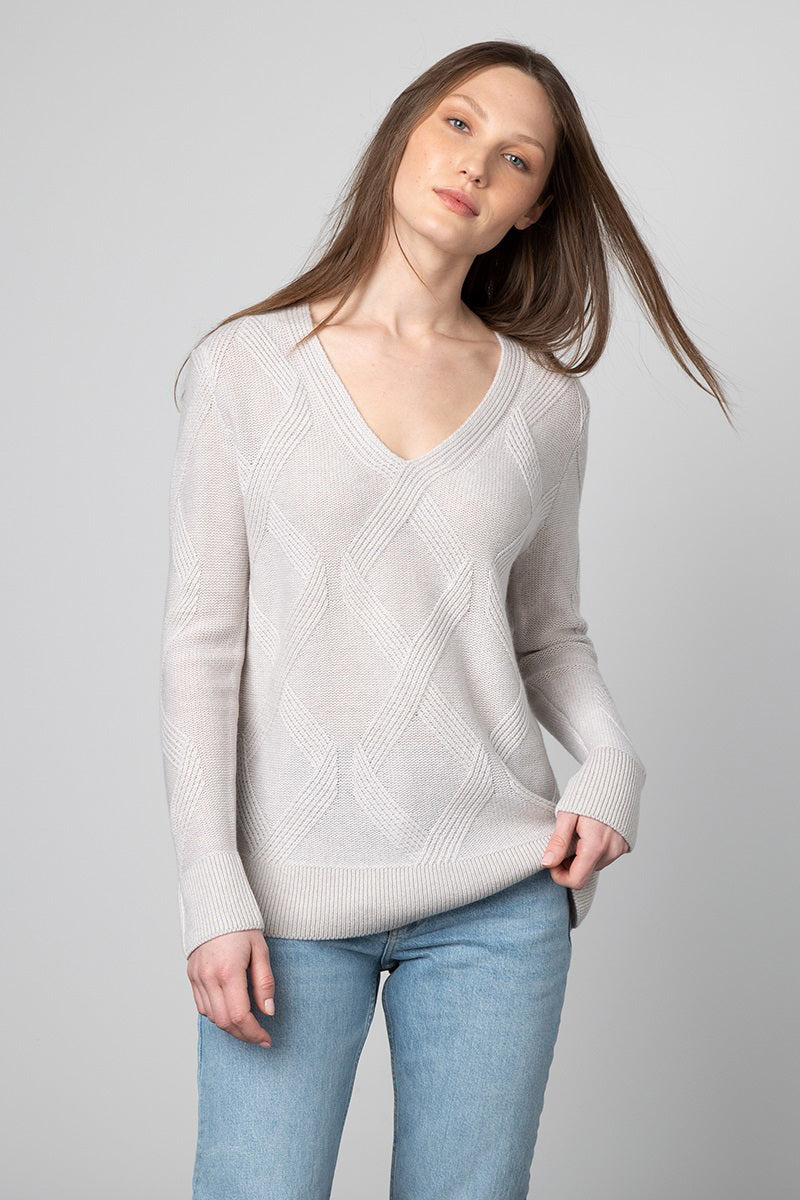 A person with long, straight hair is wearing a KINROSS CASHMERE - LUXE CABLE VEE NECK PULLOVER in light gray, paired with light blue jeans. They are standing against a plain gray background, gazing directly at the camera with a relaxed expression.