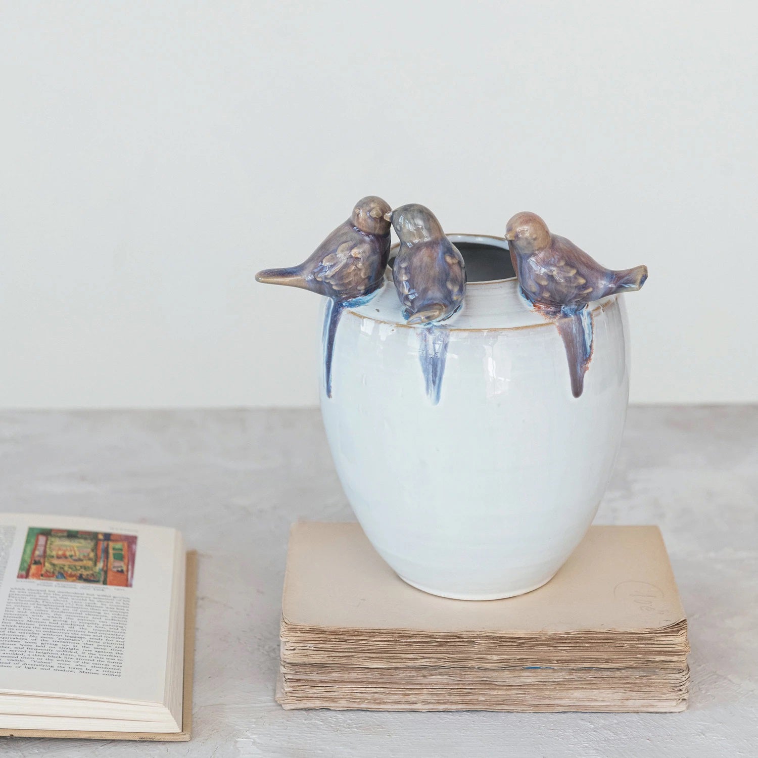 The STONEWARE PLANTER WITH BIRDS by CREATIVE COOP features a reactive glaze on white ceramic with three 3D bird sculptures on its rim, elegantly resting on two vintage books against a soft, neutral backdrop.