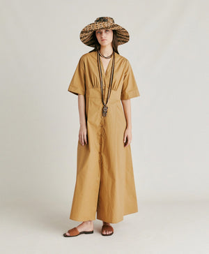 A woman stands in profile wearing the MOMONI - MARTINELLA DRESS, a long, tan cotton poplin dress from MOMONI with short sleeves and a flared skirt. She pairs it with a wide-brimmed, patterned hat, brown sandals, and a necklace. She faces left against a plain, light background.