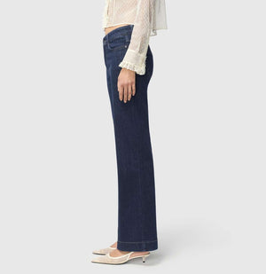 A person with light brown hair stands against a plain background, wearing a sheer, ruffled, long-sleeved white blouse and MAC JEANS - DREAM WIDE AUTHENTIC by MAC in dark blue. They're paired with comfortable white shoes.