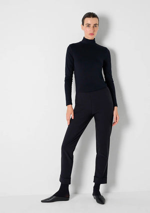 A person in KATHARINA HOVMAN - SIMPLE PANTS, featuring an elastic waistband and a black turtleneck, poses against a white wall. With short dark hair, one hand on their hip, they complete the look with sleek black footwear.