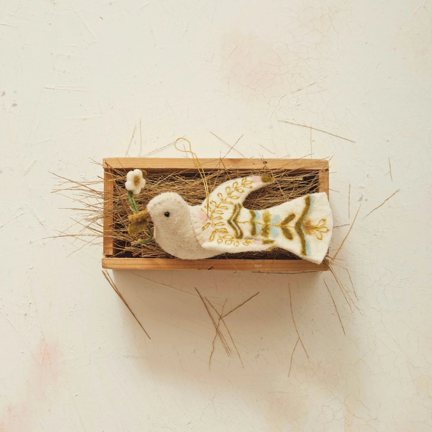 The CREATIVE COOP HANDMADE WOOL FELT DOVE ORNAMENT, featuring detailed yellow embroidery, is nestled in a small wooden box filled with straw. This charming dove holds a gentle white flower in its beak and is displayed against a light, textured background.