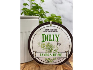 A round label for "LAMBS & THYME - DILLY DIP" featuring an illustration of dill and the text "LAMBS & THYME" is displayed in the foreground. The label emphasizes that it is free from MSG, gluten, and GMOs. In the background, there is a white container with a parsley plant growing out of it.