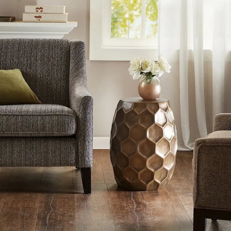 A cozy living room has a gray armchair and the OLLIIX ANTIQUE BRONZE ACCENT TABLE, which supports a vase of white flowers. A green pillow is on the couch, while sunlight filters through a curtain, brightening the wooden floor.