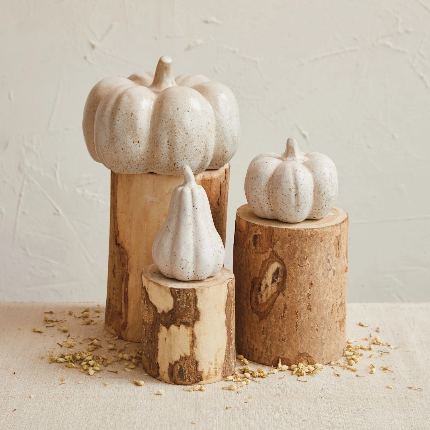 A trio of white speckled stoneware pumpkins from the Creative Coop brand, featuring various sizes, is beautifully arranged on wooden logs of differing heights. This delightful home decor piece rests on a beige surface adorned with scattered wood shavings and is set against a textured, light-colored background.