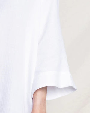 A close-up shot of a person donning the PETITE PLUME - GAUZE SHORT CAFTAN by PETITE PLUME features the loose-fitting white design with wide sleeves. The image reveals only a section of the caftan and part of their arm against a simple, light backdrop, allowing the textured and soft nature of its lightweight cotton gauze fabric to stand out.