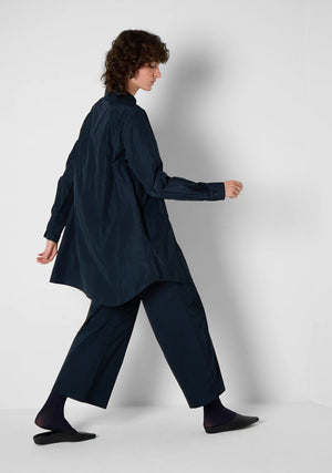 Against a light gray background, a person models the KATHARINA HOVMAN - CLASSIC OVERSIZED SHIRT with mother-of-pearl buttons and black pants. Their hair is tied back as they face away, slightly raising their arms to highlight the shirt's loose fit.