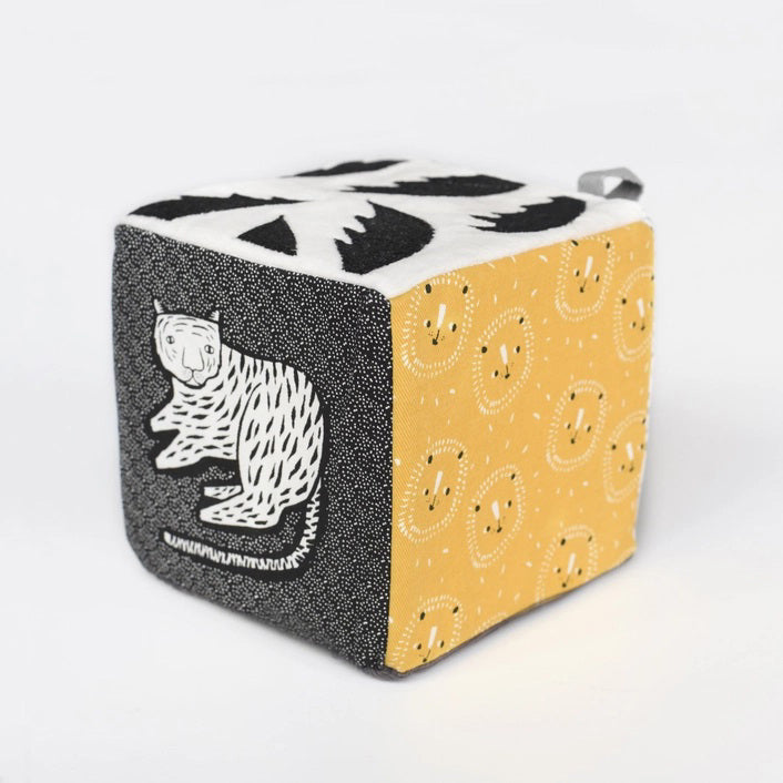 The WEE GALLERY - JUNGLE SOFT BLOCK by WEE GALLERY is a soft fabric cube perfect for stimulating a baby&#39;s developing senses. It features monochrome patterns and playful designs, including a cute monkey illustration on one side and a yellow leaf on another. Inspired by jungle life and animals, this toy boasts various textures and small loops on top for easy grasping.
