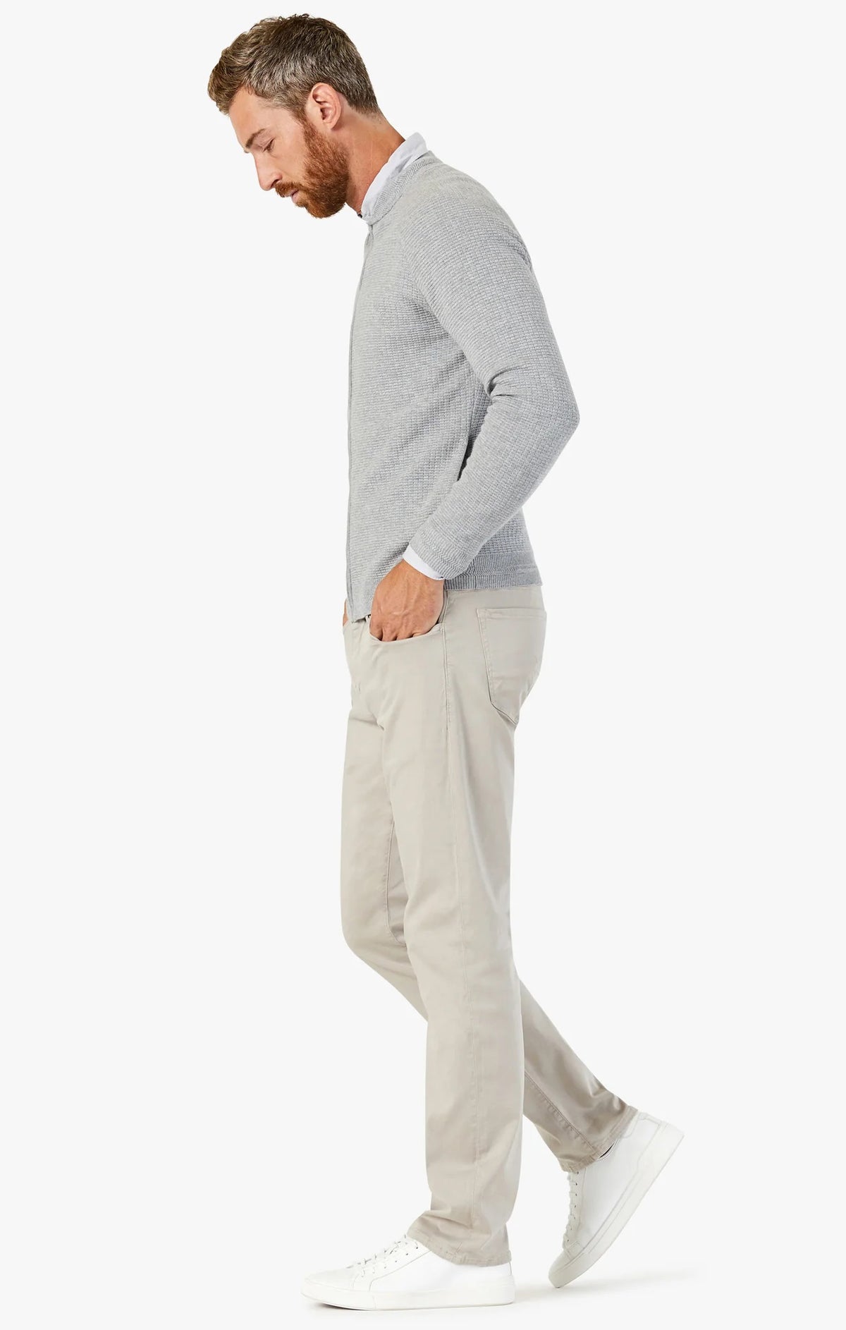 A man with short brown hair and a beard wears a light gray sweater, white sneakers, and 34 HERITAGE - COURAGE STRAIGHT LEG PANTS IN DAWN TWILL. He stands with his back to the camera, turning his head left against a plain white background.
