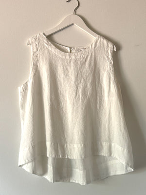 The CUT LOOSE - HI LOW TANK, a sleeveless white linen blouse with a round neckline, is displayed on a white hanger against a light gray wall. Perfect for summer wear, this breathable top by CUT LOOSE features a loose fit and an elegant high-low hem that is shorter in the front and longer in the back.