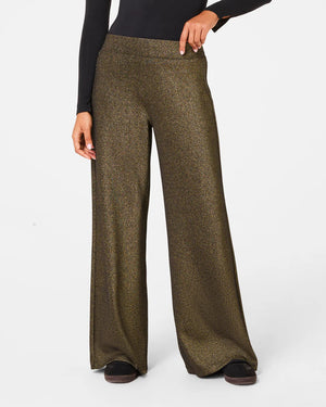 A person is wearing the SPANX AIRESSENTIALS WIDE LEG SHIMMER PANT in black along with a zip-up top. They stand casually with their hair tied back, sporting black and white sneakers, all set against a plain white background.