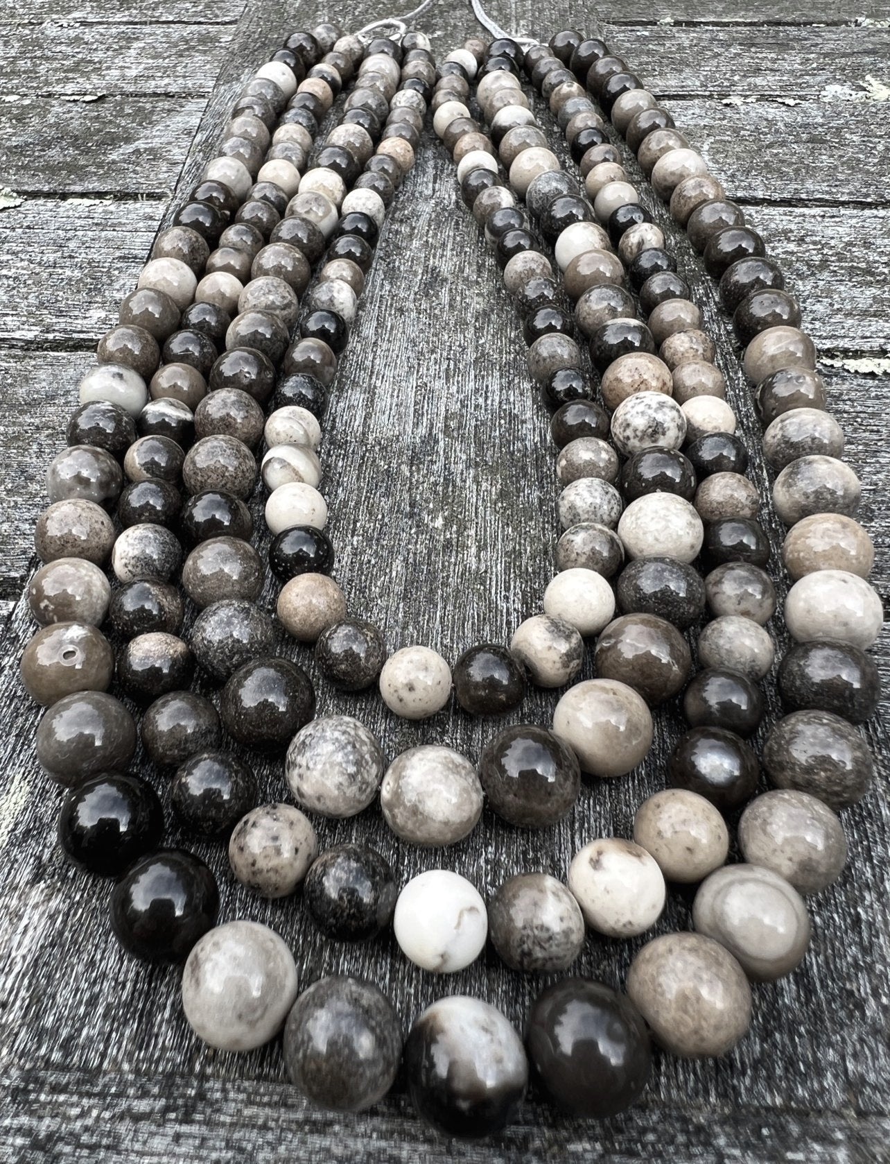 The ANN LIGHTFOOT - FOUR STRAND SHEEP SKIN JASPER NECKLACE by ANN LIGHTFOOT features a collection of multistrand necklaces composed of round, polished stone beads in varying shades of black, gray, brown, and white. These are paired with adjustable nylon ties for a custom fit. When laid out on a weathered wooden surface, the beads exhibit natural patterns and textures that evoke the beauty of sheep skin jasper.