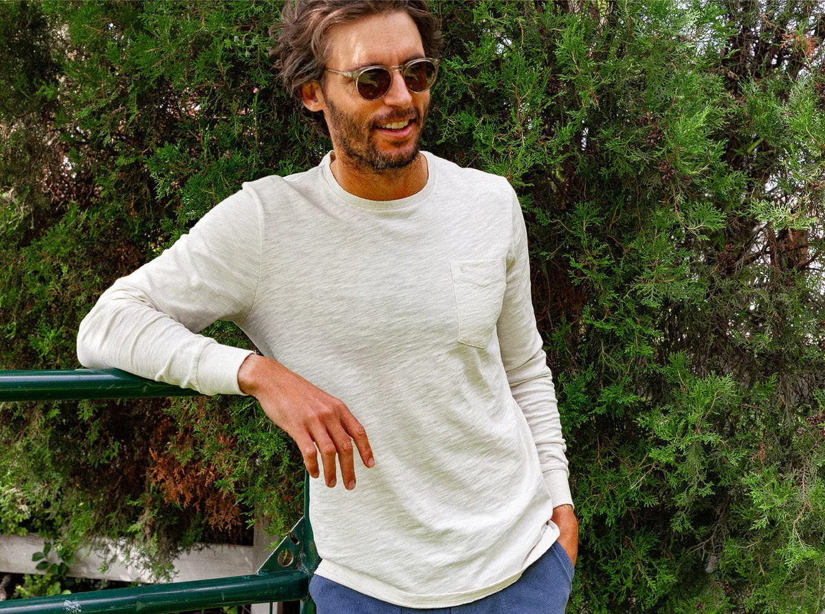 A man with short, curly hair and a beard wears the CRIQUET Cotton Slub Long Sleeve T-Shirt in Natural, styled with blue pants, standing against a plain white background.