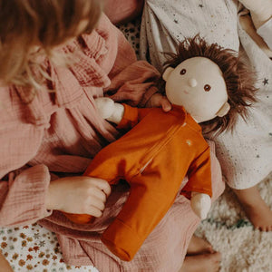 A young child in a pink outfit holds a plush doll adorned in OLLI ELLA USA's DINKUM DOLL PJS, crafted from 100% cotton. The doll features shaggy brown hair and a stitched smile. Nearby, another child is partially visible, sitting on a textured white and patterned carpet while wearing star-patterned pajamas, creating the perfect slumber party scene.