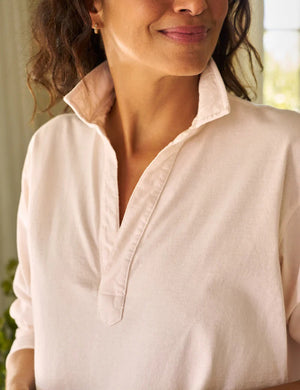 A person in a Frank & Eileen Patrick Popover Henley in Petal smiles with the collar popped, emphasizing the relaxed fit of the Heritage Jersey. The softly blurred background hints it's indoors.