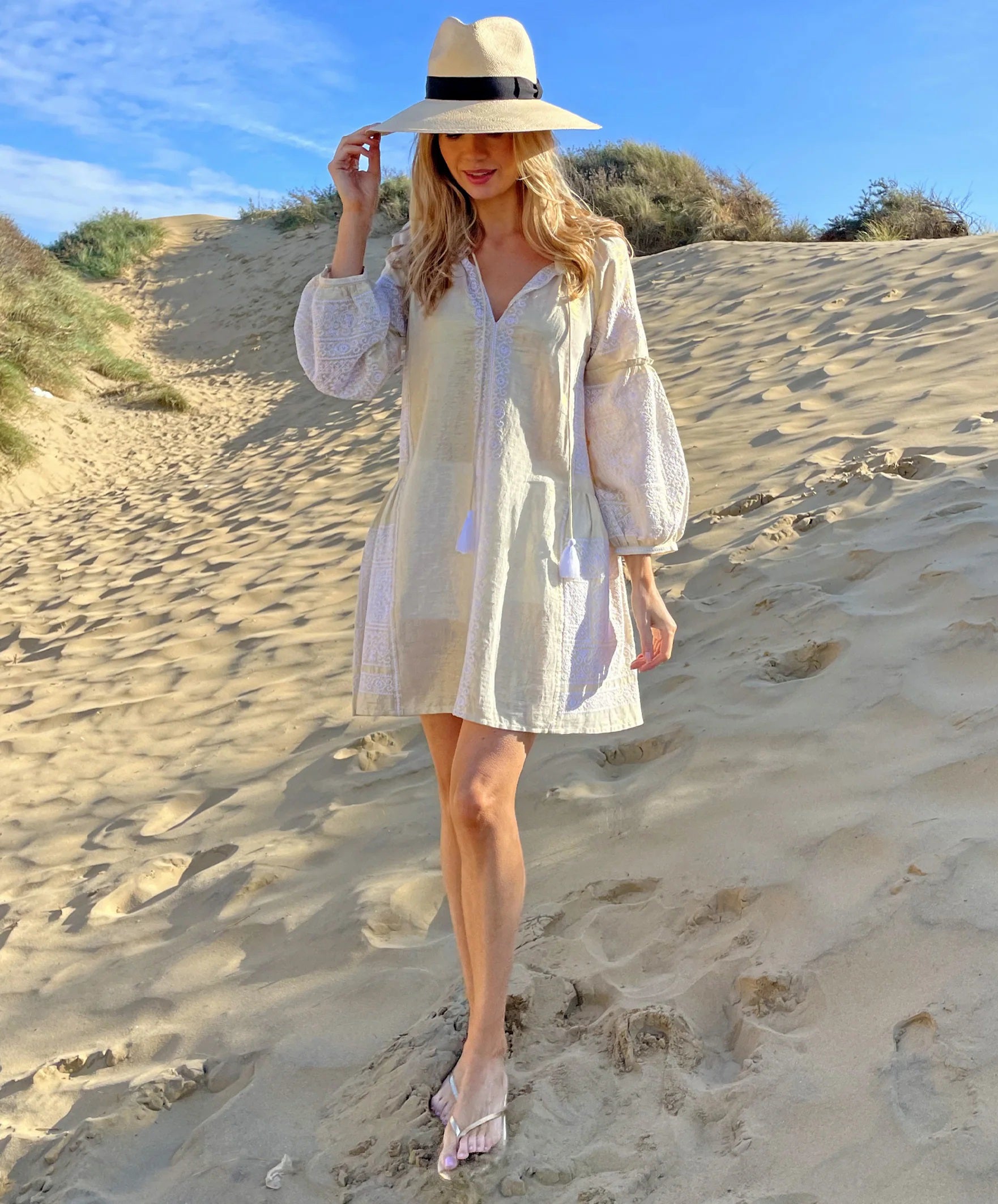 A woman stands in front of a white wall adorned with green vines and plants. She is wearing the ROSE AND ROSE - COMO DRESS from the brand ROSE & ROSE, a light-colored, long-sleeve garment with a subtle pattern, featuring delicate Cogolin knot embroidery. Her hair is down, and she is smiling as sunlight brightens the scene, casting soft shadows.