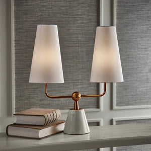 The MADISON DUBLET LAMP by NAPA HOME AND GARDEN showcases a sleek, contemporary design with a sophisticated white, conical marble base and two elegant white cylindrical lampshades. The lamp is supported by a gold metallic bar that gracefully splits into two upward arms featuring a brass finish, embodying modern elegance.