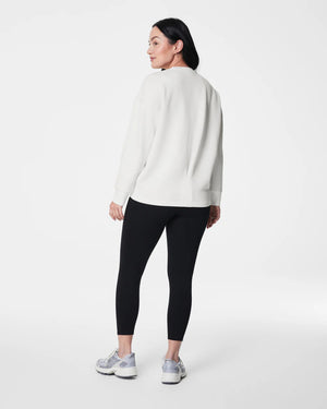 A person with long black hair stands facing slightly away from the camera, dressed in a SPANX AireEssentials Long Drew Sweater made of soft spacer fabric, complemented by black leggings and gray sneakers. The high-low hem on the sweater adds a touch of style against the plain white studio backdrop.