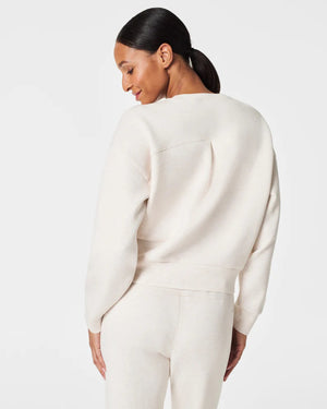 A woman stands confidently against a plain white background, wearing the SPANX Aireessentials Crew Neck Pullover and matching sweatpants made from luxurious comfort spacer fabric. She has her hair pulled back and her left hand is raised slightly. She smiles gently with her lips closed.