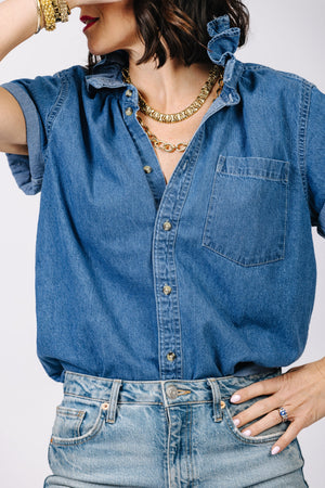 An individual is sporting the KMJ - QUARANTINI SHORT SLEEVE SHIRT, featuring a ruffle collar, rolled sleeves, and a front pocket, styled with layered gold necklaces and multiple bracelets. The person is also wearing denim jeans and has white nail polish. The person's face is partially out of frame.