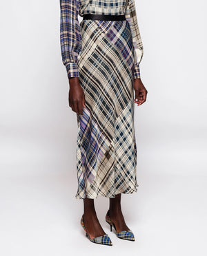 A person is modeling an uncomplicated chic outfit that includes the MIRTO 1956 - BLUE PLAID MIDI SKIRT paired with a long-sleeved, high-necked blouse, pointed heels, and holding a small, dark handbag. The skirt features a cinched waist and long design, and the person has short hair.