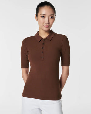 A woman in the SPANX - STRETCH RIB POLO TOP, a brown, ribbed, short-sleeve shirt with a button-down front, stands with her arms relaxed. The plain white background accentuates the top's stretchy ribbed fabric and body-hugging fit.