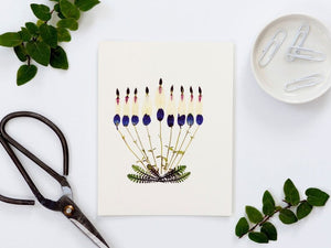 An elegant design of botanical arrangements featuring assorted dried flower petals, leaves, and small plant parts decorates the white background of the HAPPY HANUKKAH HOLIDAY CARD from PETAL PEOPLE PRESS, complete with a pair of tweezers and scissors.