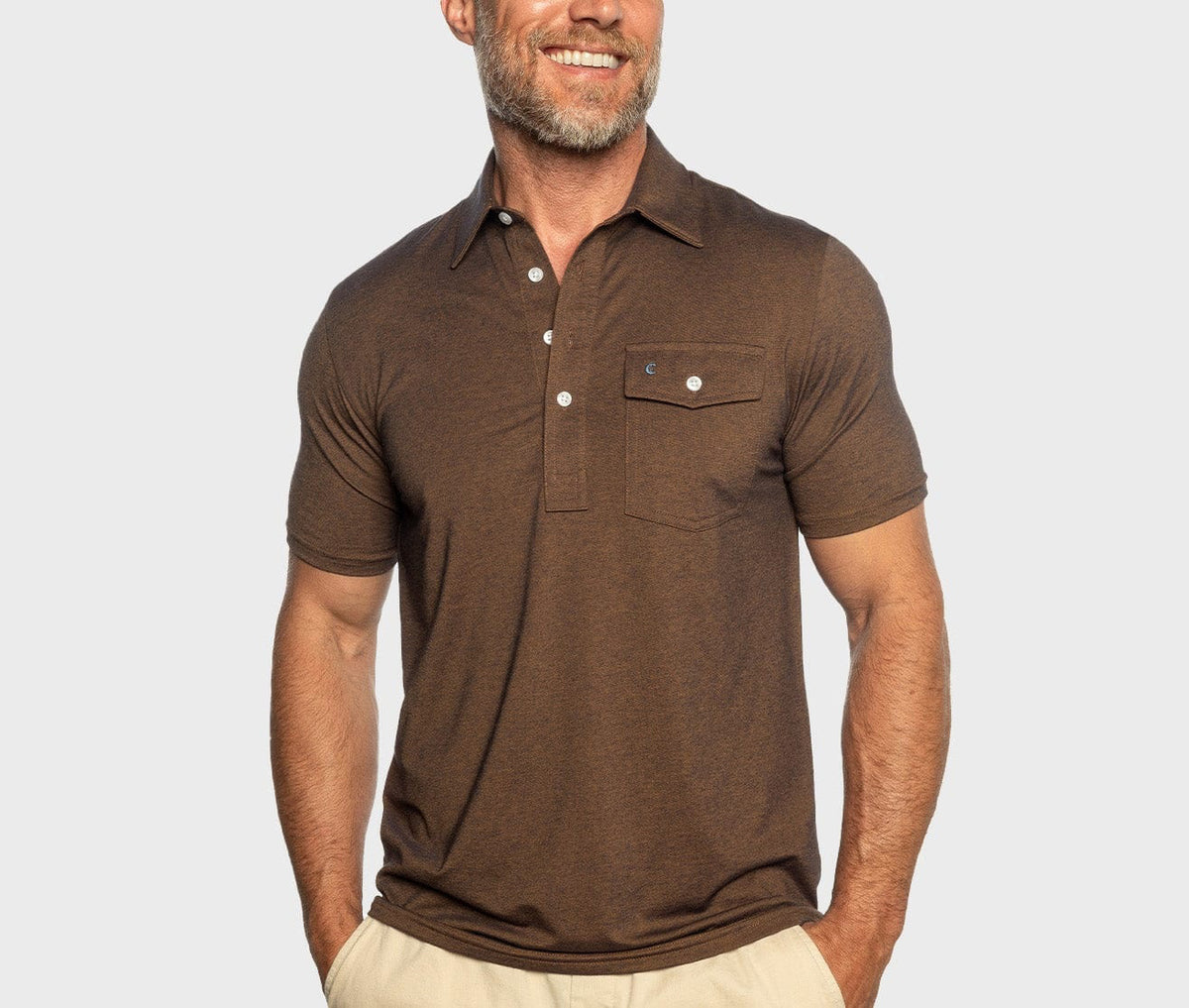A smiling man wears the CRIQUET Performance Players Shirt in WoodsMoke—a short-sleeved brown shirt with a buttoned chest pocket—paired with cream pants, standing against a light gray background.