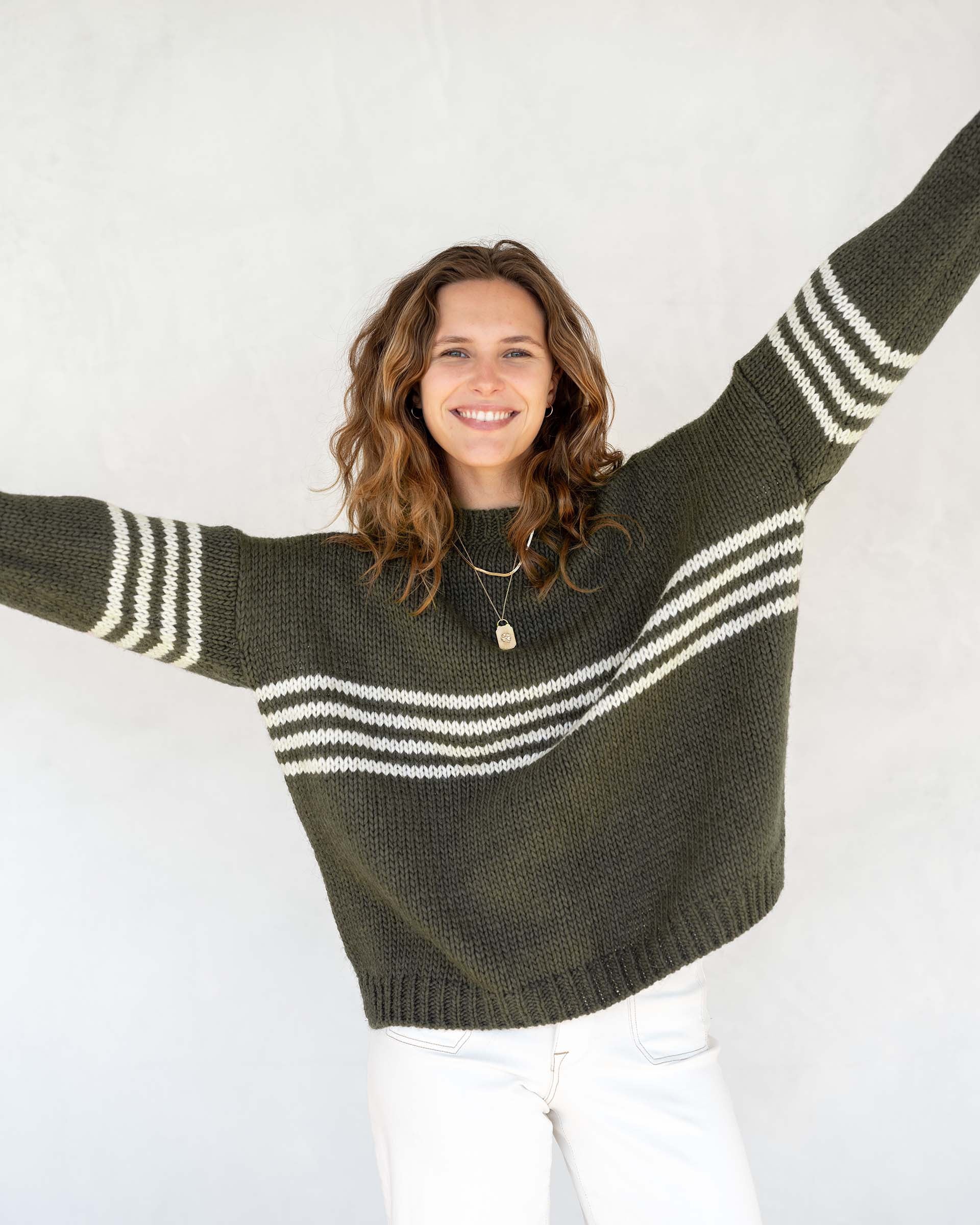 A person with long, wavy brown hair is wearing the MERSEA - SEACOAST STRIPE SWEATER, a loose-fitting, cozy blue knit sweater with white stripes on the sleeves, paired with black pants. They are standing against a plain, light-colored background and facing left, with their hands near their face.