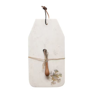 The CHEESE BOARD WITH CANAPE KNIFE by CREATIVE COOP is a rectangular white marble cheese board embellished with a leather hanging loop. It is elegantly decorated with a tied rustic wooden spoon, decorative leaves at the bottom corner, and features a stunning mother of pearl inlay that adds an exquisite touch to any kitchen or dining setting.