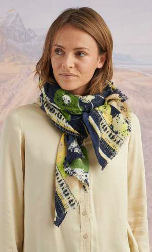 A woman outdoors with a blurred mountain backdrop wears a cream button-up and the STORIATIPIC DORIS Wool Silk Scarf by STORIATIPIC, featuring green, navy, and yellow patterns in a luxe wool-silk blend.