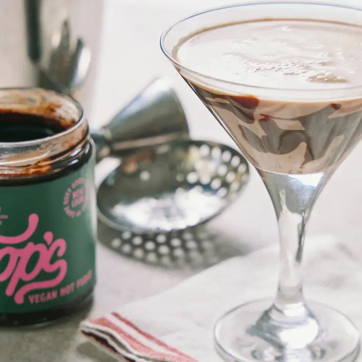 A jar of COOP'S - VEGAN HOT FUDGE with chocolate dripping from the lid. The label is green with pink cursive text and states "Smooth Rich Flavor," "High-grade European chocolate with fresh extracted coconut," and "New Look." This handmade chocolate sauce is the best ice cream topping. Net weight 10oz (283.5g).