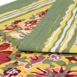 A wooden table features the Jardin Red and Green Table Runner by Couleur Nature, adorned with a lit candle, pinecones, and greenery. Red napkins and silverware are neatly placed next to a light blue bowl on the runner.