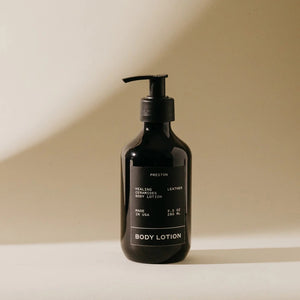 A sleek, dark brown bottle of Preston Body Lotion with a pump dispenser is displayed against a neutral beige background. The label reads "PRESTON," "Healing Amber Body Lotion," "Made in USA," and "9.5 fl oz / 266 ml." Infused with plant-based moisturizers, its natural formula is accentuated by soft lighting that casts shadows to the right of the bottle.