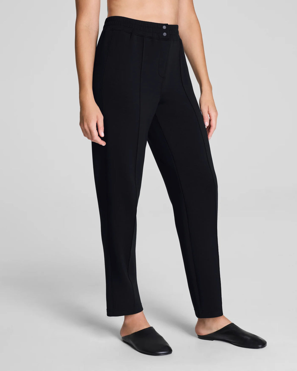 A person models the SPANX - AIRessentials Brushed Slim Straight Pant with an elastic waistband and buttons, paired with black slip-on shoes against a plain light background.