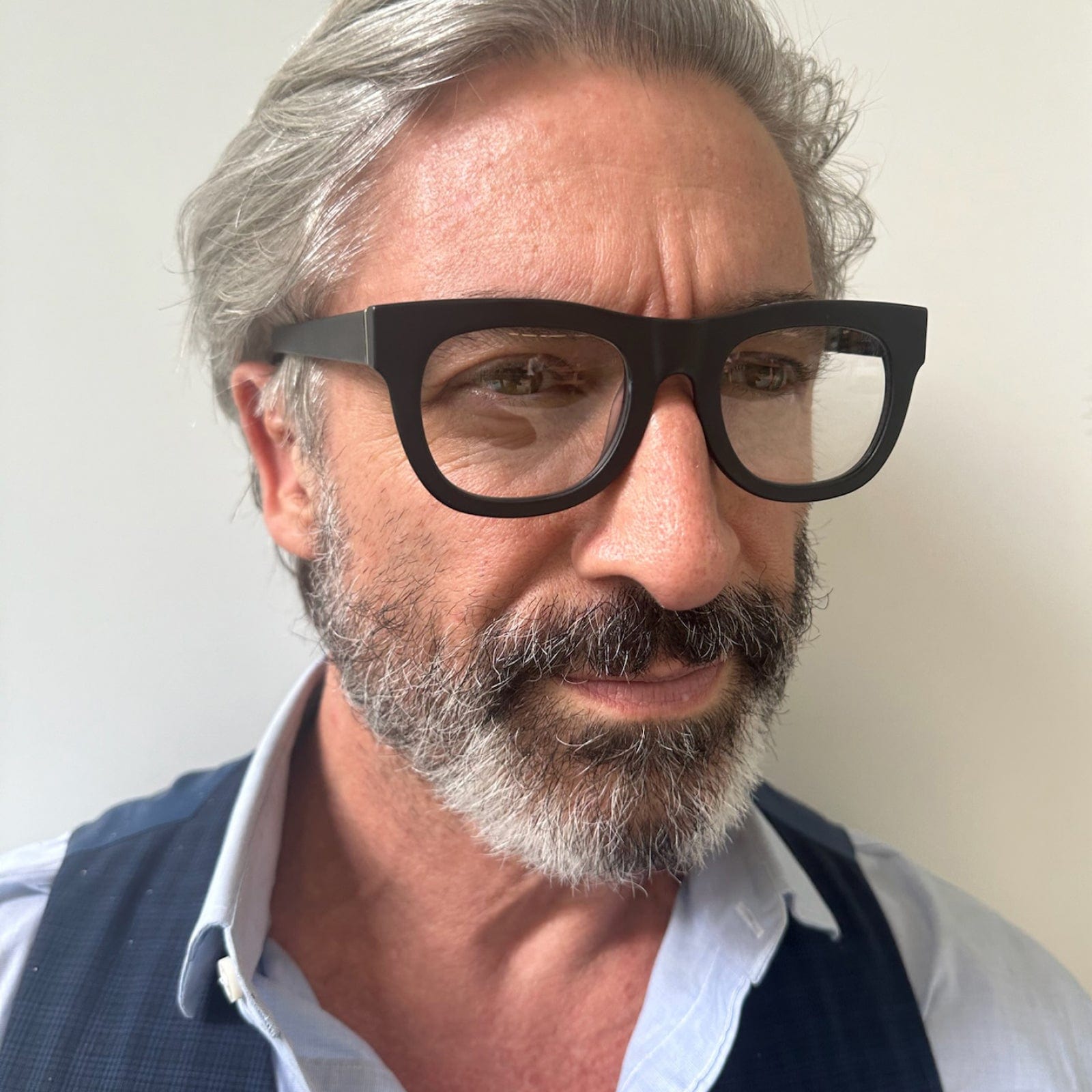 A middle-aged man with gray hair, a full gray beard, and black glasses is wearing a light blue shirt and a dark vest. He is looking slightly to the side with a neutral expression against a plain light background.