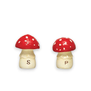 On a textured white surface, there is a serving dish shaped like a mushroom accompanied by crackers. Nearby are two hand-painted ceramic salt and pepper shakers from Creative Coop, each designed in the shape of mushrooms. One shaker features a red cap with white polka dots labeled "P" for pepper, while the other is similarly styled and labeled "S" for salt.