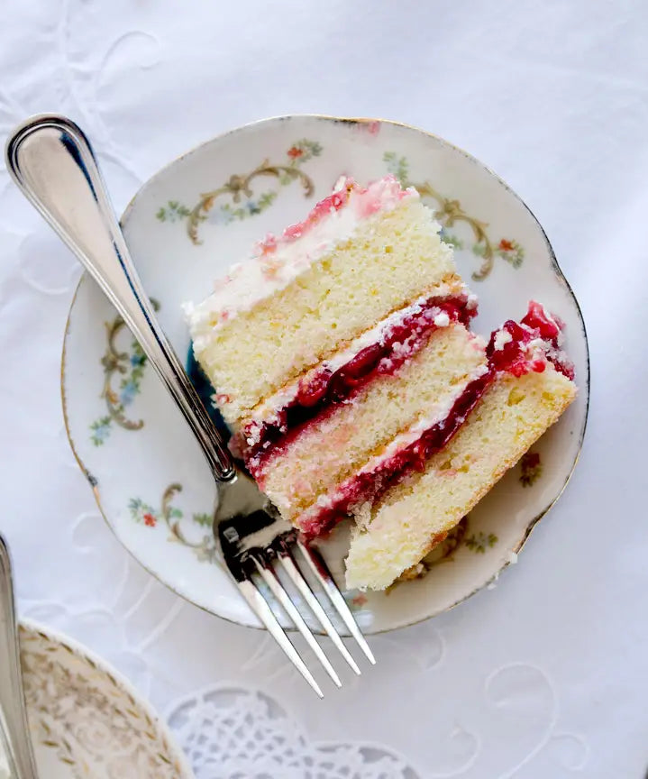 A slice of layered vanilla cake with white frosting and red fruit filling sits on a decorative floral plate with a fork beside it, on a white tablecloth.
