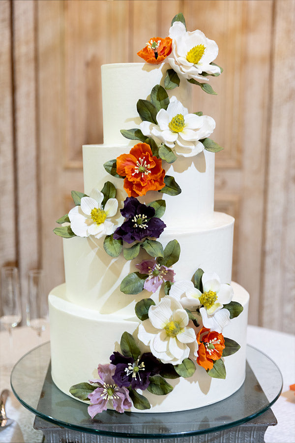 A four-tiered white cake decorated with realistic sugar flowers in white, orange, and purple, cascading diagonally with green leaves. The cake sits on a glass stand against a wooden background.