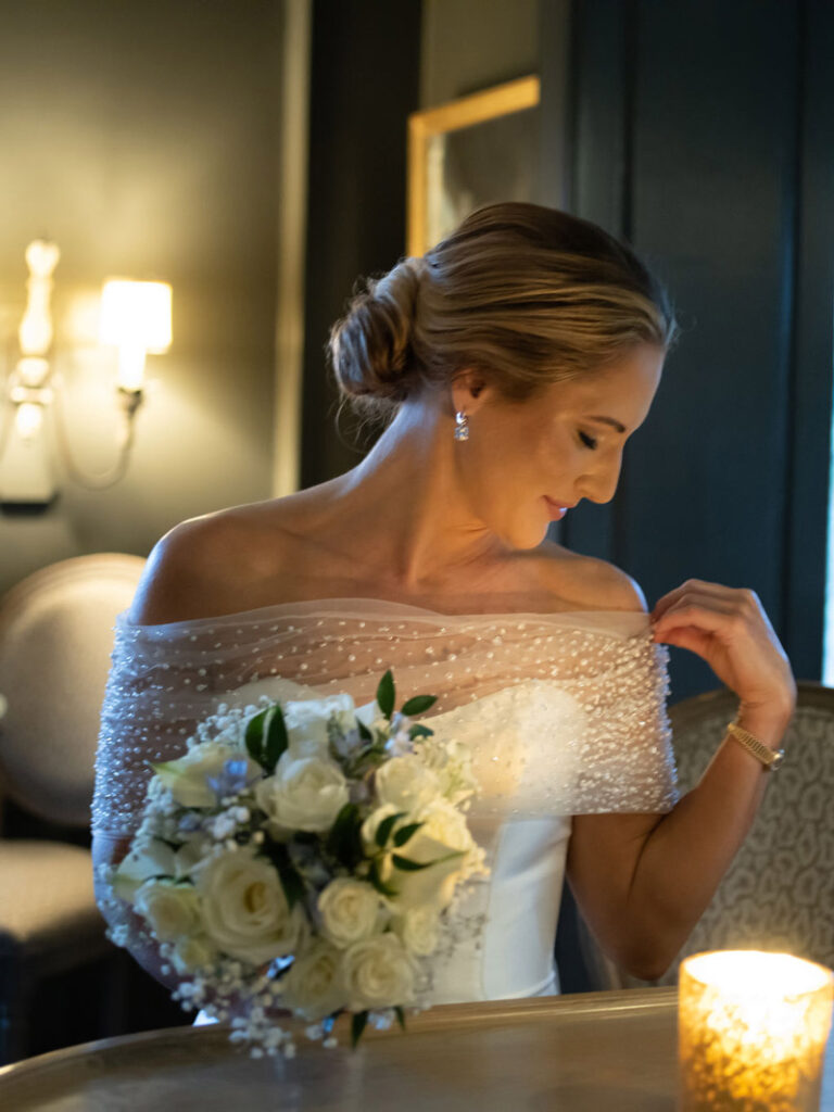A bride in an off-the-shoulder wedding dress holds a white rose bouquet and looks down, gently touching her beaded veil. The scene is softly lit by a nearby candle.