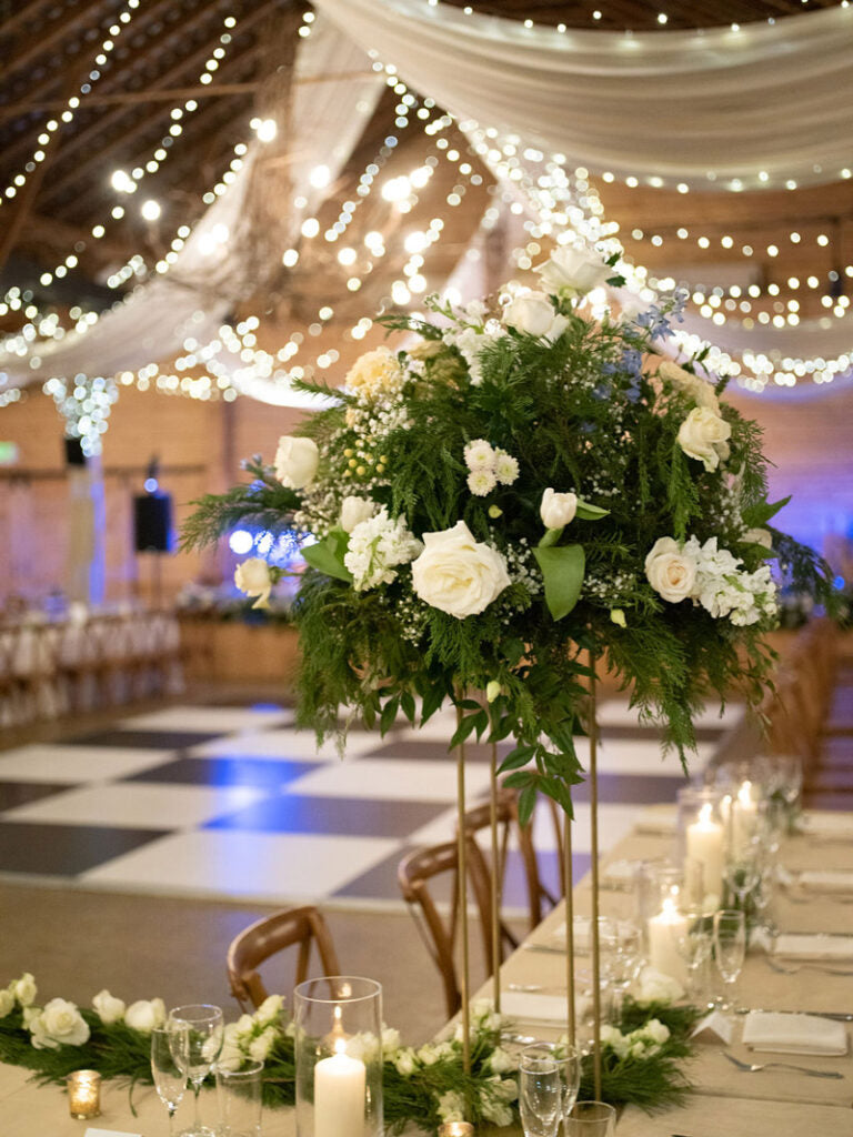 A wedding reception hall with string lights and draped fabric overhead, featuring a tall floral centerpiece with white roses and greenery on a decorated dinner table with candles and elegant table settings.