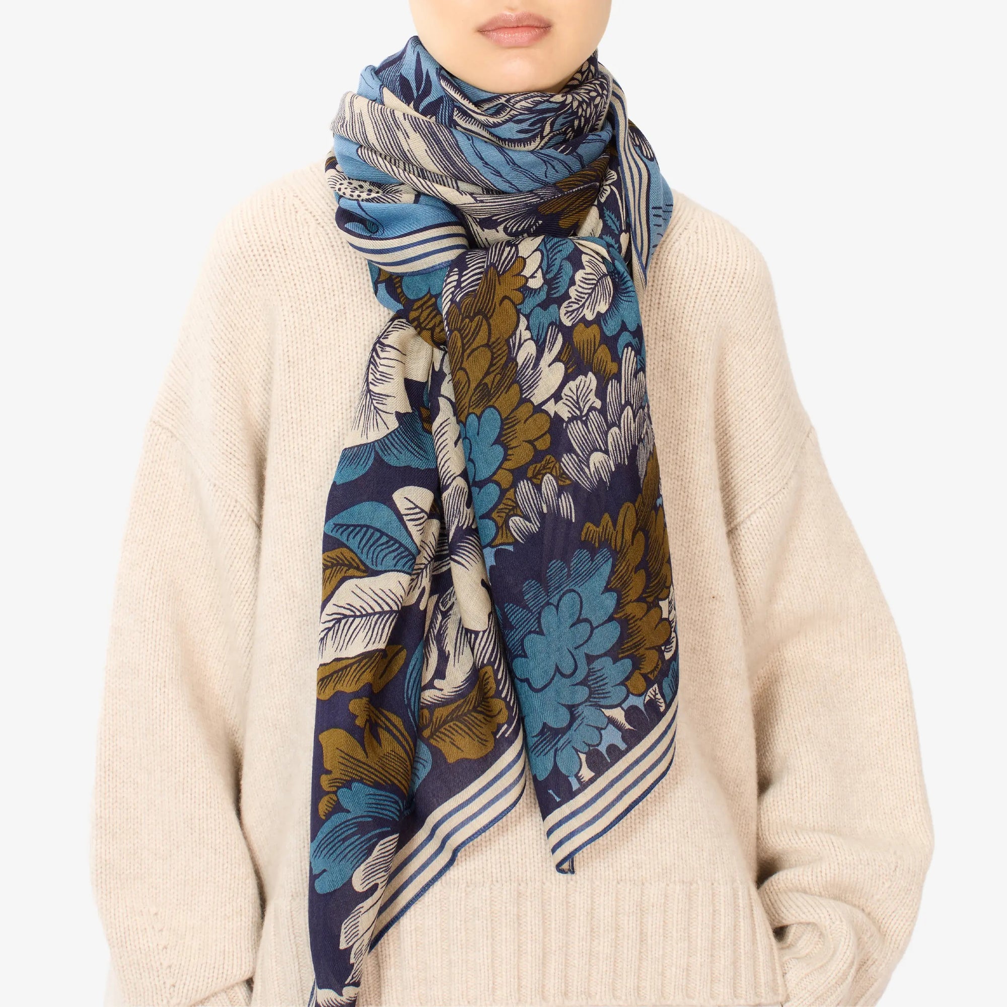 A person is wearing a beige sweater and the INOUI EDITIONS - PANTHERE SCARF, which features a blue, brown, and white floral pattern. The scarf made of washable wool is wrapped around their neck. The plain white background emphasizes the scarf and sweater, with the individual's face partially visible.