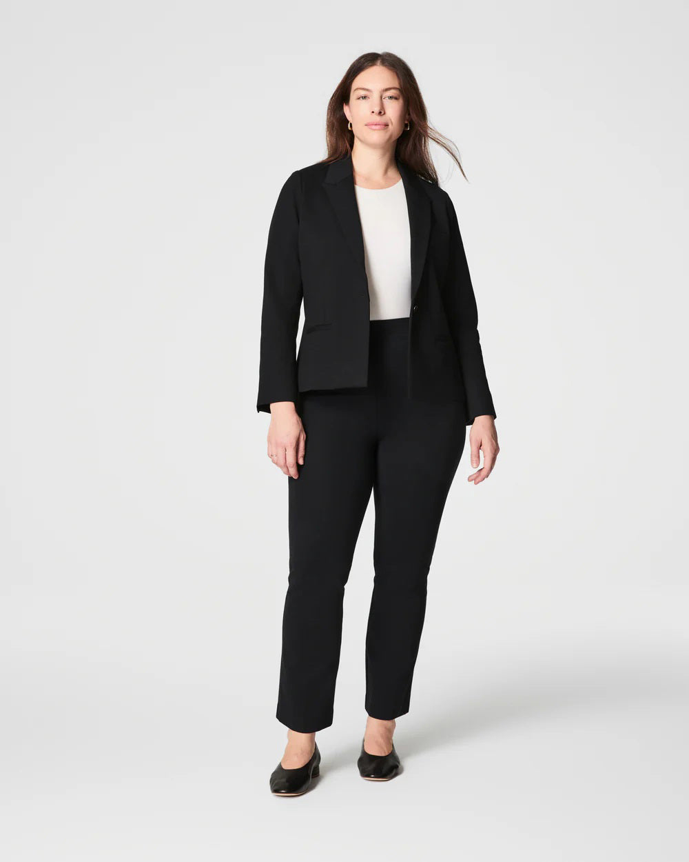 A person with long hair poses confidently, wearing the SPANX - PONTE BLAZER in a versatile black hue, crafted from premium ponte fabric, layered over a white top and dark jeans. The plain, light-colored background creates a minimalist setting.